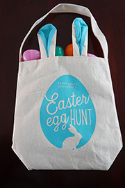 A canvas tote bag that says Saint Louis University Easter Egg Hunt with an image of an egg surrounding the words and an outline of a bunny underneath. Easter eggs can be seen spilling out of the top of the bag.