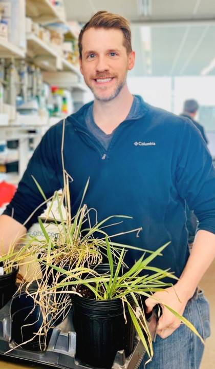 Photo of Matthew Rubin standing in a lab, holding a tray with several plants.