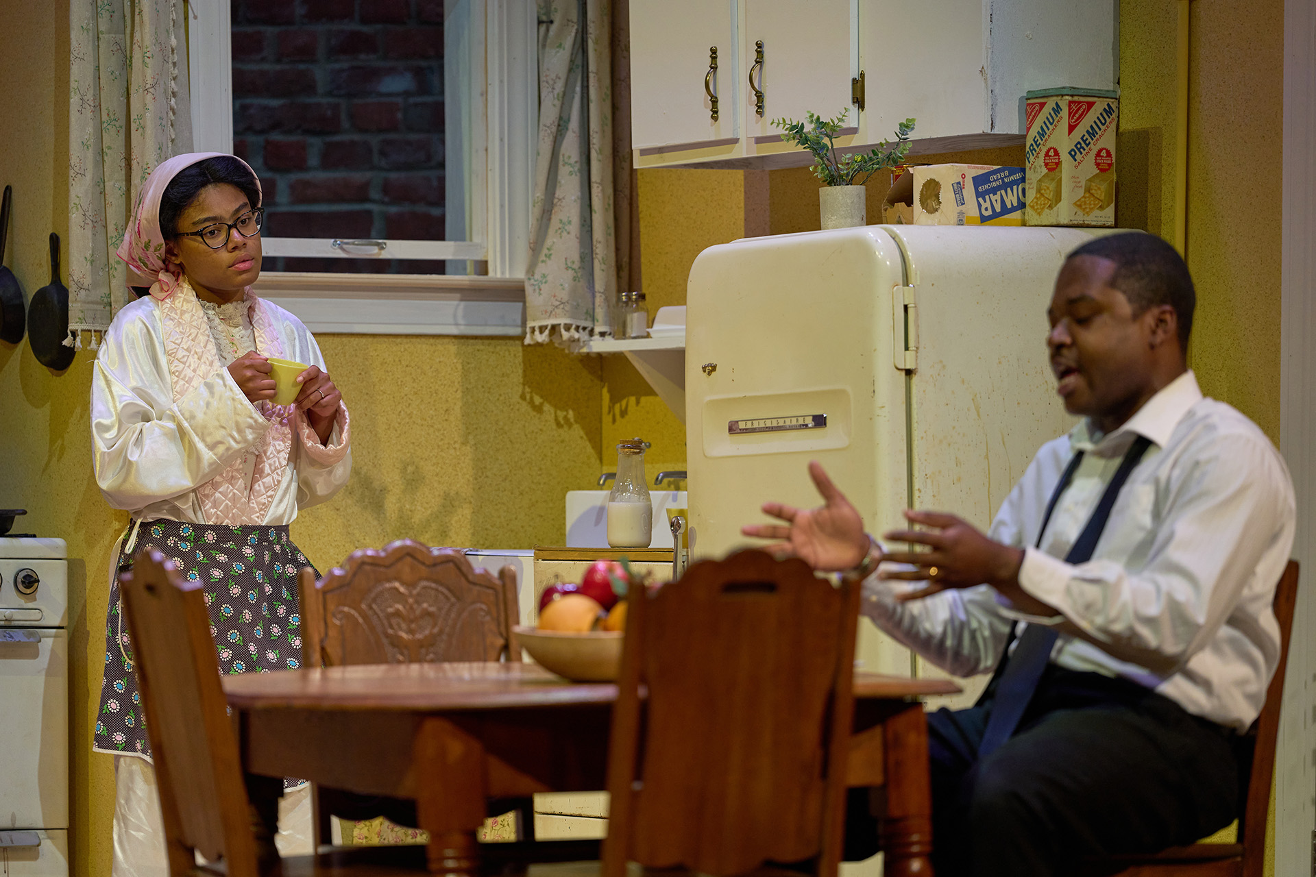 A young Black woman in houseclothes holds a teacup while looking at her husband seated at the kitchen table