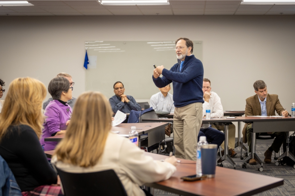 Participants seated at tables in a classroom setting, with a presenter standing in the front of the room delivering a presentation.