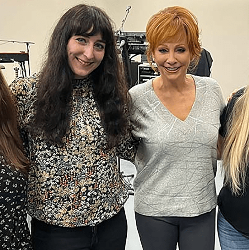 A woman in a blouse stands next to a woman in a sweater who is country music legend Reba McEntire.