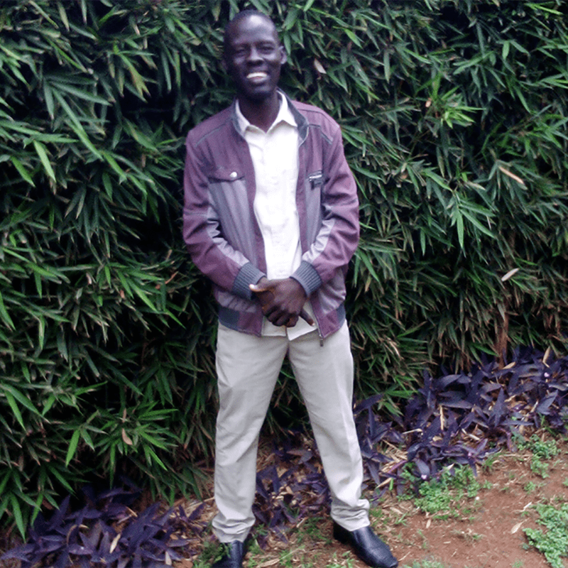 A man wearing a jacket, button-down shirt and pants stands in front of a tree and smiles.