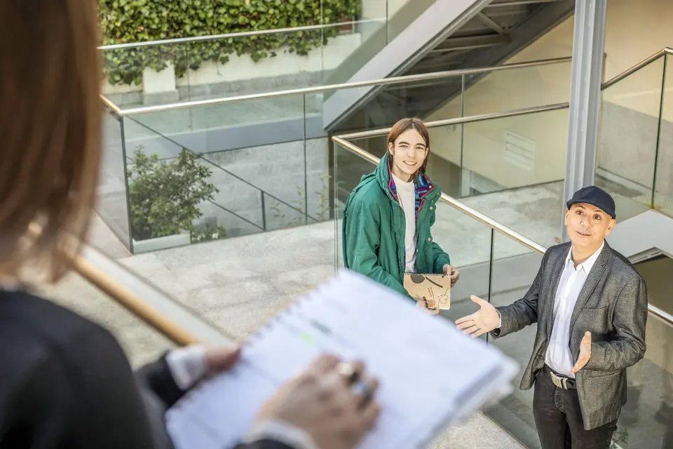 A teacher, next to a student, talks to another student holding a notebook in the foreground.