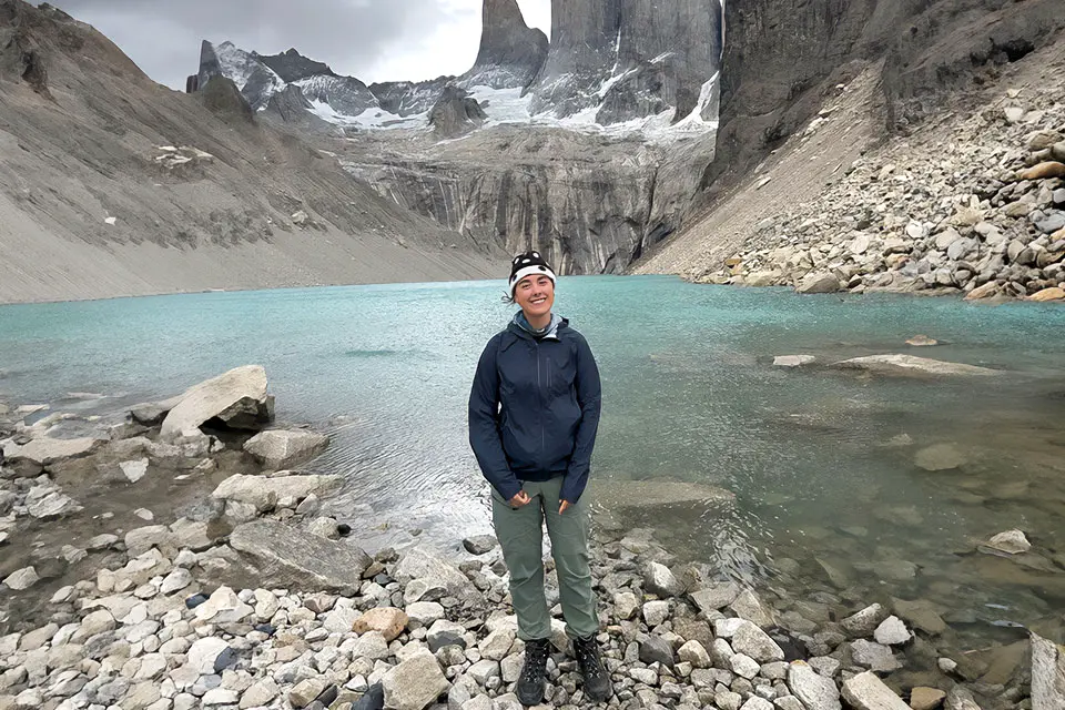 A student wearing hiking gear stands on a rocky shoreline next to a body of water, surrounded by rocky cliffs and mountains.