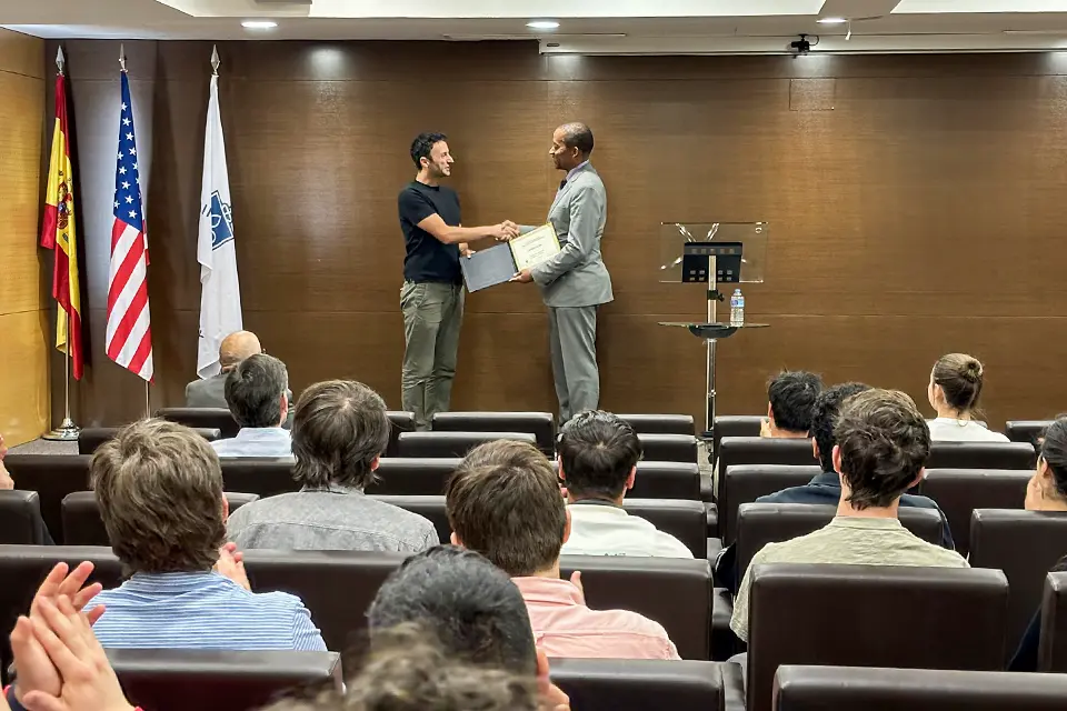 A person holding a diploma standing on a stage in front of a group of people hands the diploma to the winner.