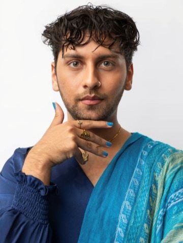 Anuj Gandhi Anuj Gandhi poses for a photo against a white background. Anuj rests his right hand under his chin. Anuj wears a blue and teal multi-patterned long-sleeve shirt.