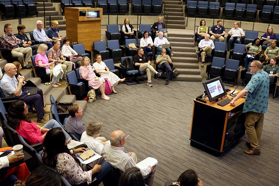 A man addresses an audience from a podium inside a classroom with stadium seating. 