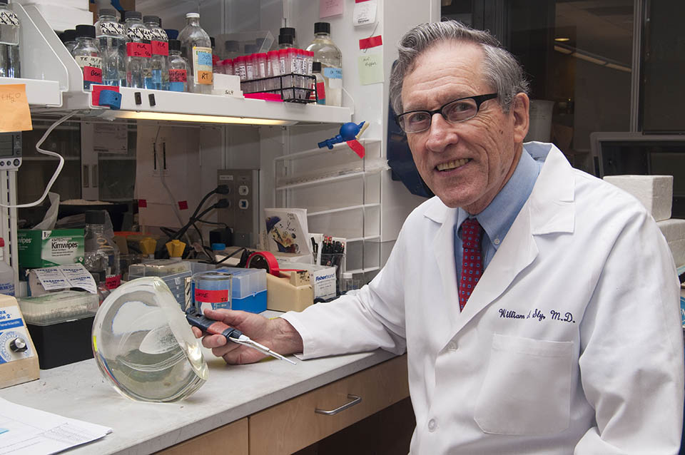 Dr. Sly smiles for a photo in a lab. He is wearing a white lab coat and a long instrument in his right hand which rests on a countertop. Glass bottles and vials with red tops are on a top shelf. 