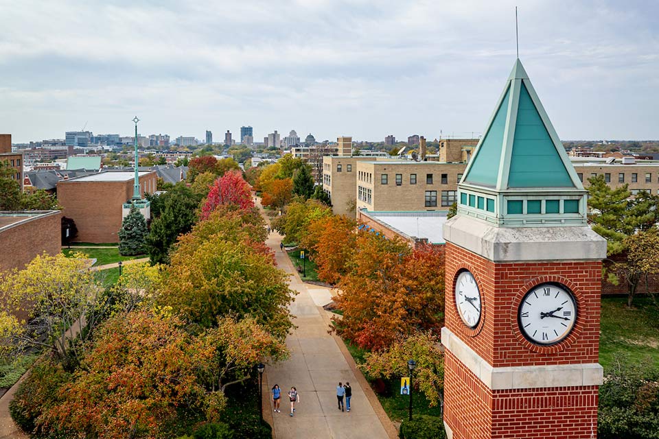 Aerial view of a college campus in autumn, featuring a brick clock tower with teal accents. Colorful trees line the walkway with distant city skyline.