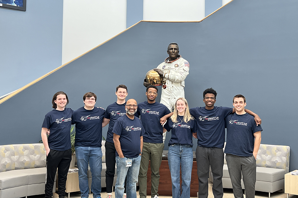 Several people pose for a photo in front of a sculpture of an astronaut in a white suit holding a gold helmet inside a building.t