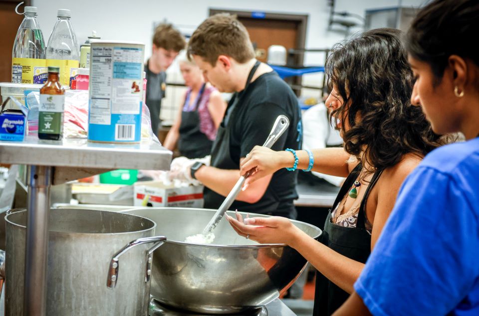 A student wearing a blue t-shirt watches as a student stirs a stainless steel bowl in a kitchen. 