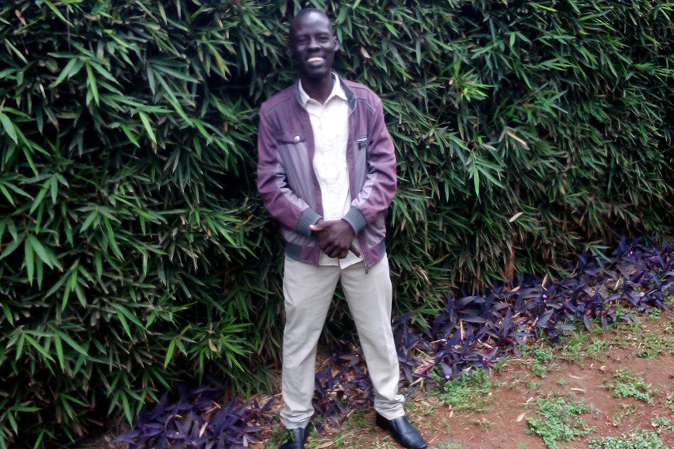 A male student poses for a photo outdoors. He wears a maroon jacket, white button-up shirt and beige slacks. He smiles for the camera. 