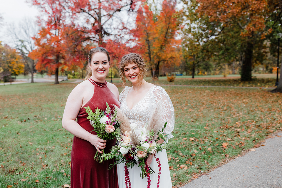 Two women dressed in wedding attire and holding a bouquet of flowers smile for a photo in a park on a fall, cloudy day. 