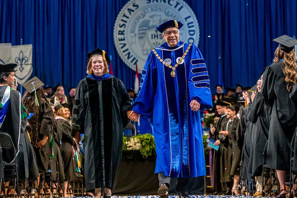 President Fred P. Pestello and his wife, Fran, leave a commencement ceremony walking hand-in-hand. Dr. Pestello wears a blue gown and hat with a gold tassel and chain. Fran wears a black gown with a gold tassel.