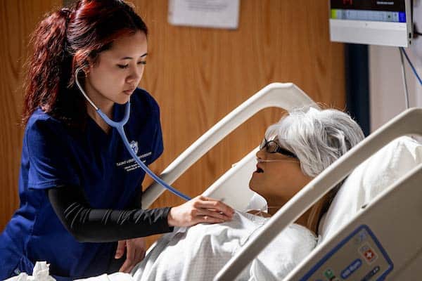 A female student wearing SLU scrubs examines an adult manikin lying in a hospital bed. She is using a stethoscope and using it to listen to the manikin’s heartbeat