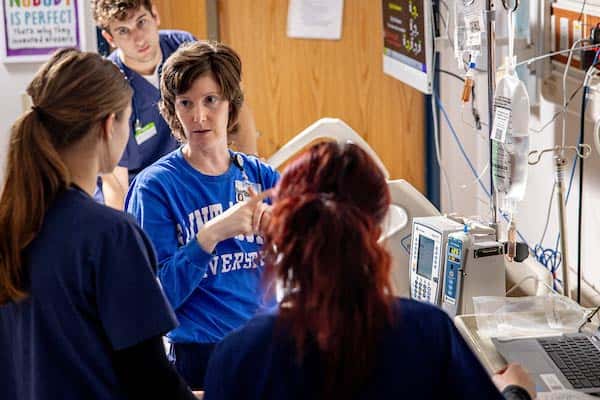 A female professor, wearing a blue Saint Louis University sweatshirt, speaks to a group of nursing students in a simulation lab. She is standing near clinical equipment placed near a hospital bed.