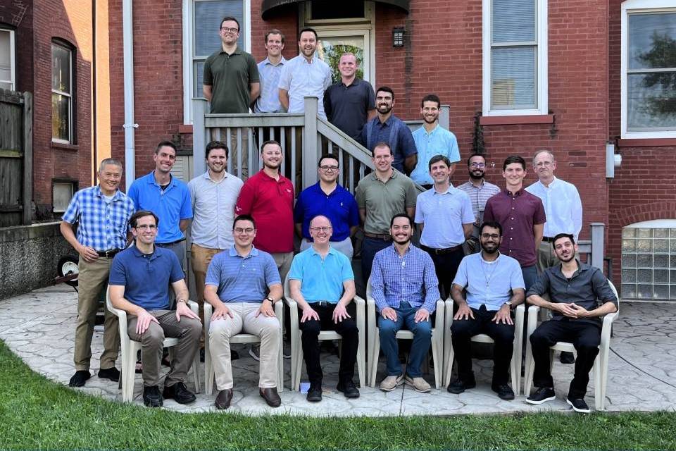 A group of residents pose for a photo in the back of the Jesuit house, some sitting on lawn chairs, and some standing in a row behind and on steps.