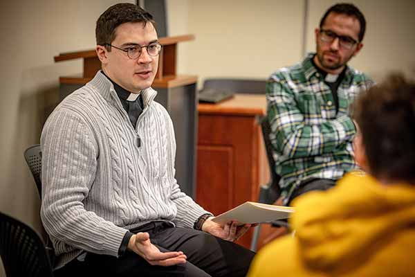 A young Jesuit wearing a priest’s collar leads a circle discussion in a classroom.