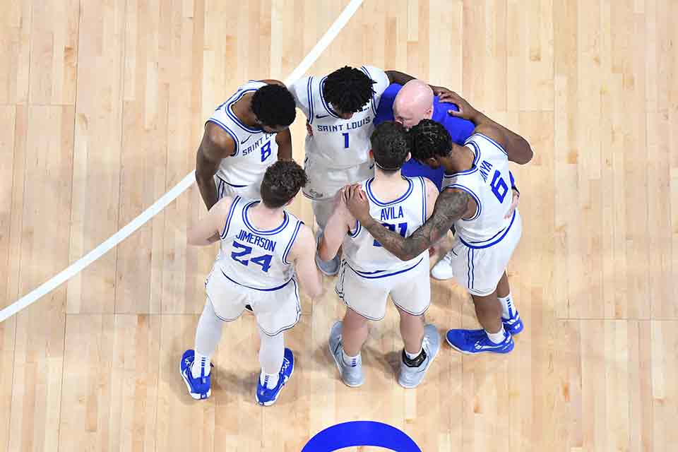 Members of the men's basketball team stand in a huddle in the middle of the court during a game.