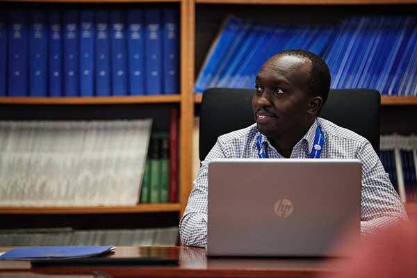 A student in a checkered button up shirt sits at a computer with a SLU lanyard in front of a bookshelf of blue books.