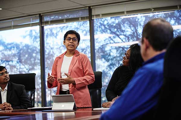 A student in a pink suit coat presents to a faculty member at a conference table.