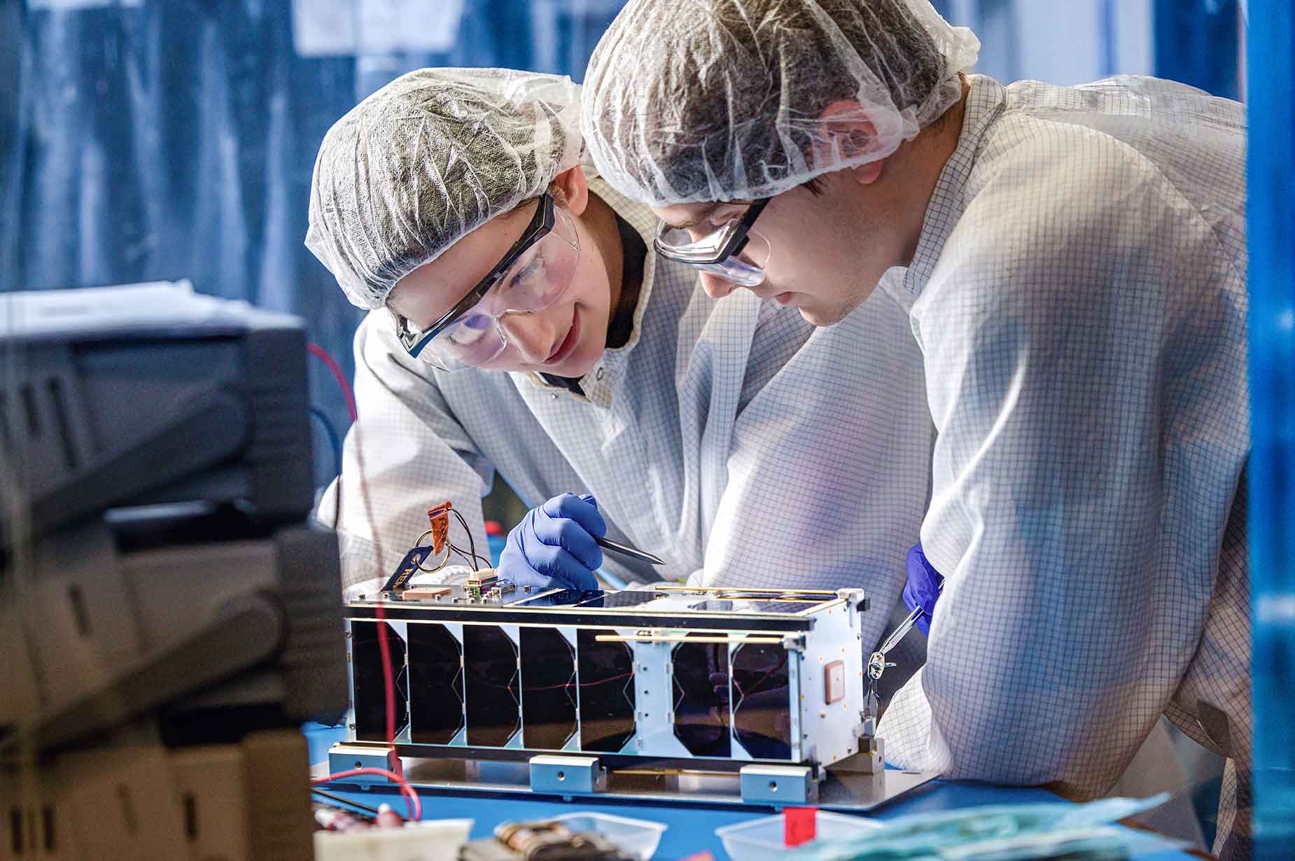Two students in personal protective equipment examine a rectangular device while standing at a lab desk.