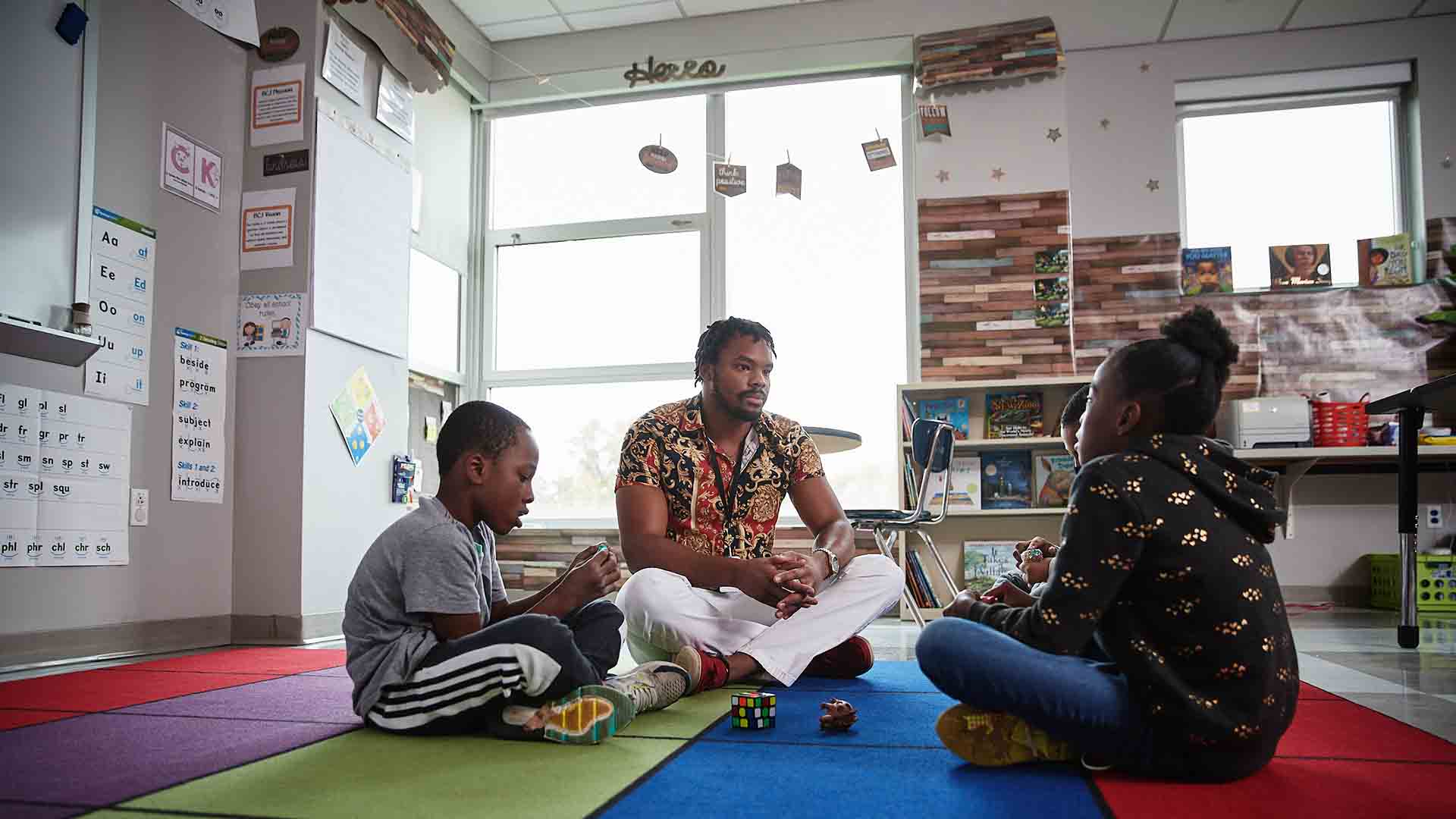 A School of Social Work student sits on the floor of a colorful classroom, talking to two young children.